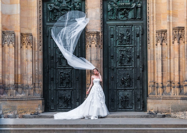 Žena v dlhých bielych svadobných šatách vychádza z kostola a vo vzduchu jej veje závoj.jpg
